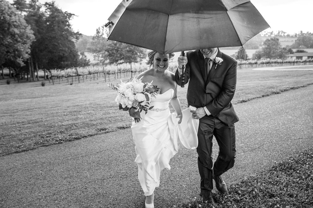 bride and groom running towards the venue from the vineyards with an umbrella over their head at Arrington Vineyards