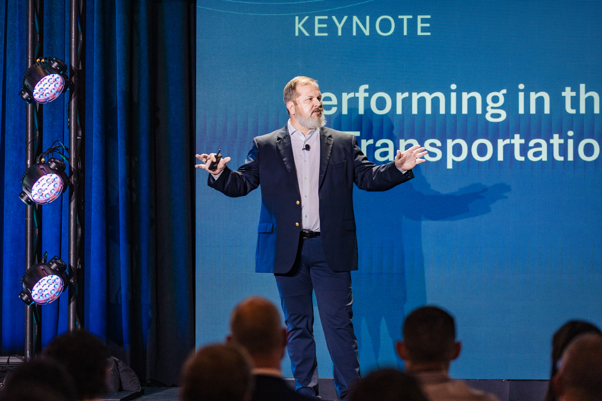 A man in a suit stands on a stage and talks to a large crowd in a conference room.